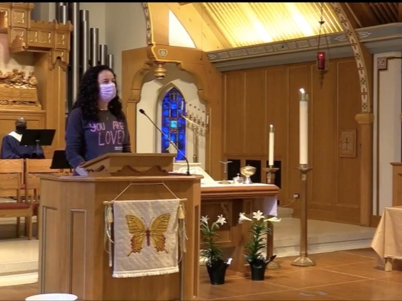 Nicole standing at lectern in church sanctuary wearing mask, shirt says 